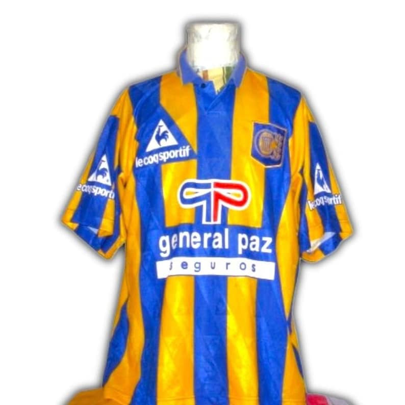 May include: A vintage soccer jersey with vertical stripes of yellow and blue. The jersey features the text "general paz seguros" and the logo of "le coq sportif". The jersey has a blue collar and short sleeves.