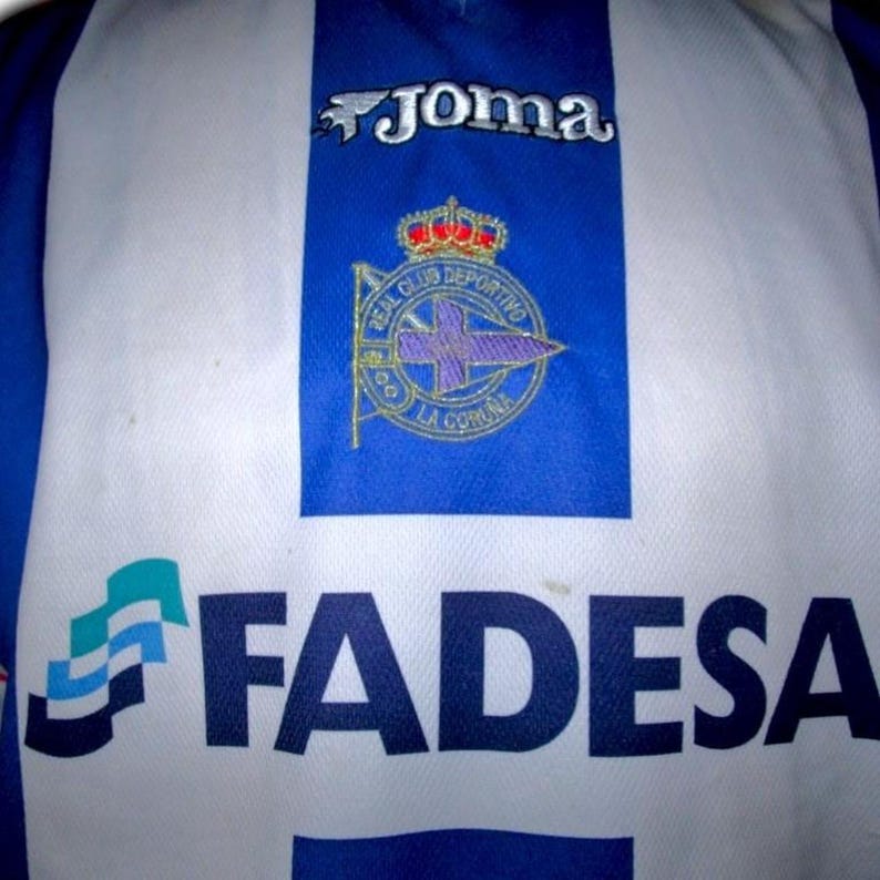 May include: A blue and white soccer jersey with the "Joma" logo at the top. The jersey features the Real Club Deportivo La Coru&ntilde;a crest and the "FADESA" sponsor logo.