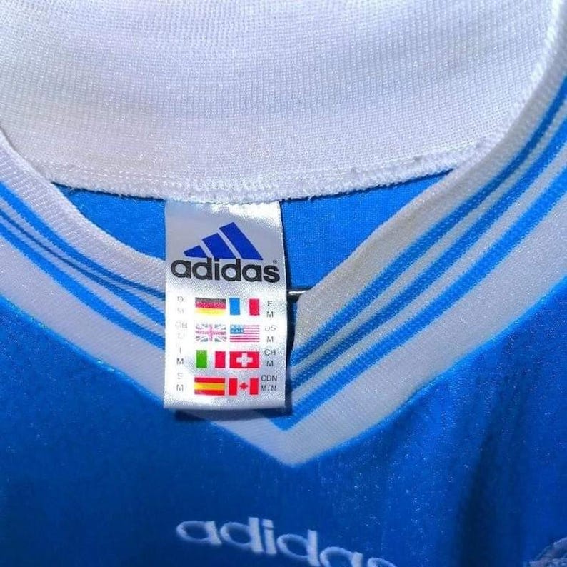 Puede incluir: Primer plano de una camiseta Adidas azul con rayas blancas y cuello blanco. El logotipo de Adidas es visible en una etiqueta con varias banderas de pa&iacute;ses. El logotipo de Adidas tambi&eacute;n es visible en la parte inferior de la camiseta.