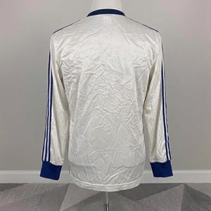 K&ouml;nnte beinhalten: Wei&szlig;es Langarmshirt mit marineblauen Akzenten. Das Shirt hat marineblaue Streifen an den &Auml;rmeln und marineblaue B&uuml;ndchen. Der Kragen ist ebenfalls marineblau. Das Shirt wird auf einer Schaufensterpuppe pr&auml;sentiert.