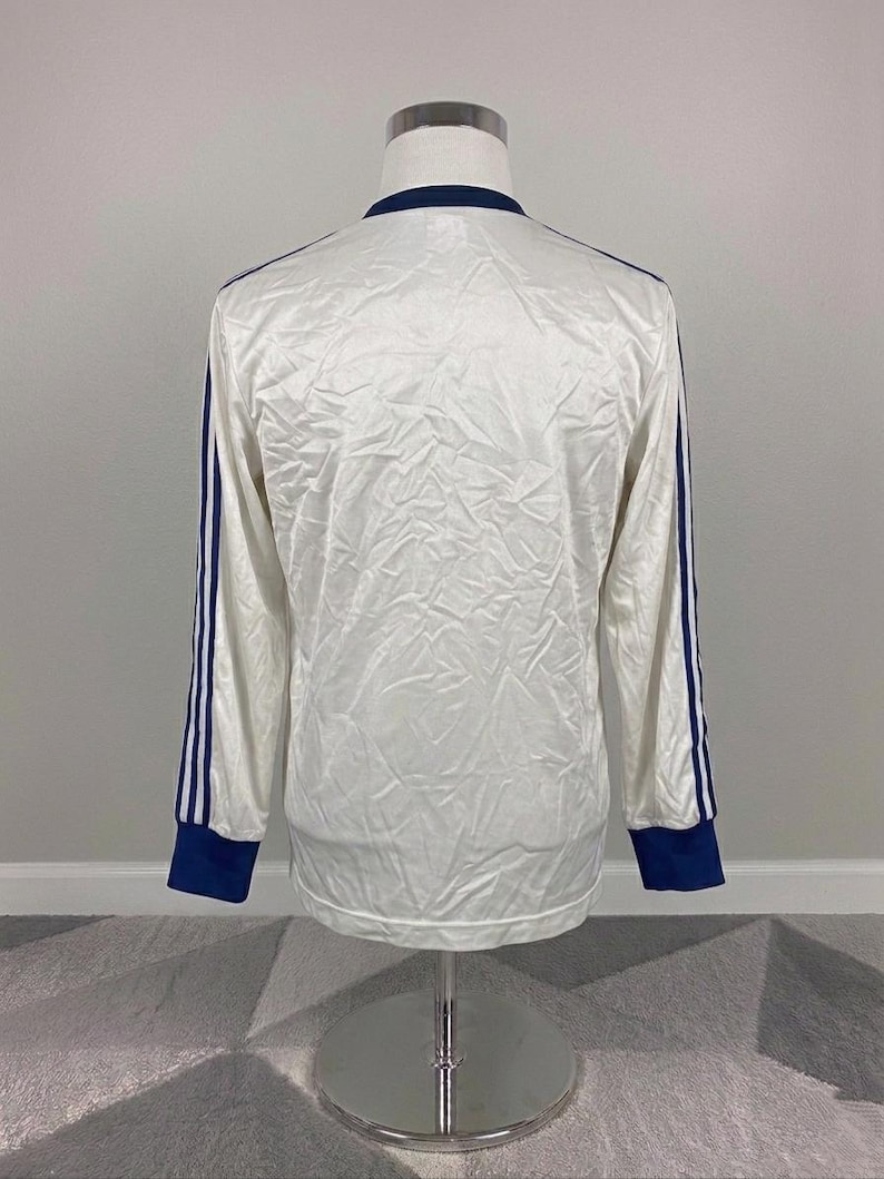 K&ouml;nnte beinhalten: Wei&szlig;es Langarmshirt mit marineblauen Akzenten. Das Shirt hat marineblaue Streifen an den &Auml;rmeln und marineblaue B&uuml;ndchen. Der Kragen ist ebenfalls marineblau. Das Shirt wird auf einer Schaufensterpuppe pr&auml;sentiert.