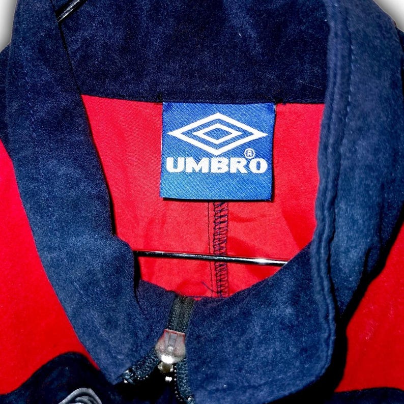 K&ouml;nnte beinhalten: Nahaufnahme einer roten und marineblauen Umbro-Jacke. Die Jacke hat einen marineblauen Kragen und Rei&szlig;verschluss, mit dem Umbro-Logo auf einem blauen Etikett. Das Innere der Jacke ist rot.