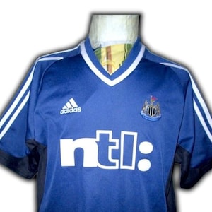May include: Blue soccer jersey with white stripes on the shoulders and sleeves. Features the Adidas logo, a team crest, and the word "ntl:" in white block letters. The jersey has a V-neck collar with white trim.