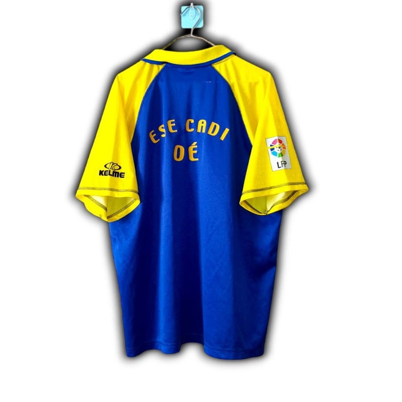 K&ouml;nnte beinhalten: Ein blau-gelbes Fu&szlig;balltrikot mit dem Text "ESE CADI O&Eacute;" in goldfarbenen Buchstaben auf dem R&uuml;cken. Das Trikot hat gelbe &Auml;rmel und einen gelben Kragen. Das Kelme-Logo befindet sich auf dem linken &Auml;rmel.