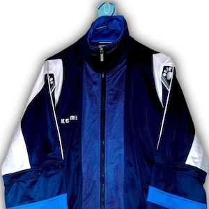 K&ouml;nnte beinhalten: Eine marineblaue und wei&szlig;e Trainingsjacke mit durchgehendem Rei&szlig;verschluss. Die Jacke hat einen hohen Kragen und wei&szlig;e Akzente an Schultern und &Auml;rmeln. Der Markenname "Keme" ist auf der Brust sichtbar.