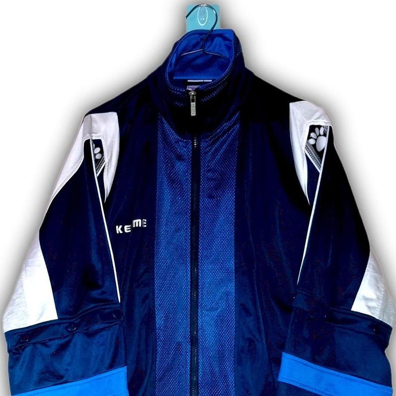 K&ouml;nnte beinhalten: Eine marineblaue und wei&szlig;e Trainingsjacke mit durchgehendem Rei&szlig;verschluss. Die Jacke hat einen hohen Kragen und wei&szlig;e Akzente an Schultern und &Auml;rmeln. Der Markenname "Keme" ist auf der Brust sichtbar.