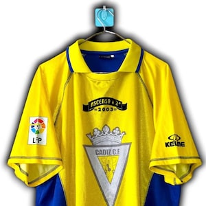 K&ouml;nnte beinhalten: Gelb-blaues Fu&szlig;balltrikot von C&aacute;diz C.F. Das Trikot tr&auml;gt den Schriftzug "ASCENSO a 2&ordf; 2003" und das Vereinswappen. Es hat auch die Logos von LFP und Kelme. Das Trikot h&auml;ngt an einem blauen Haken.