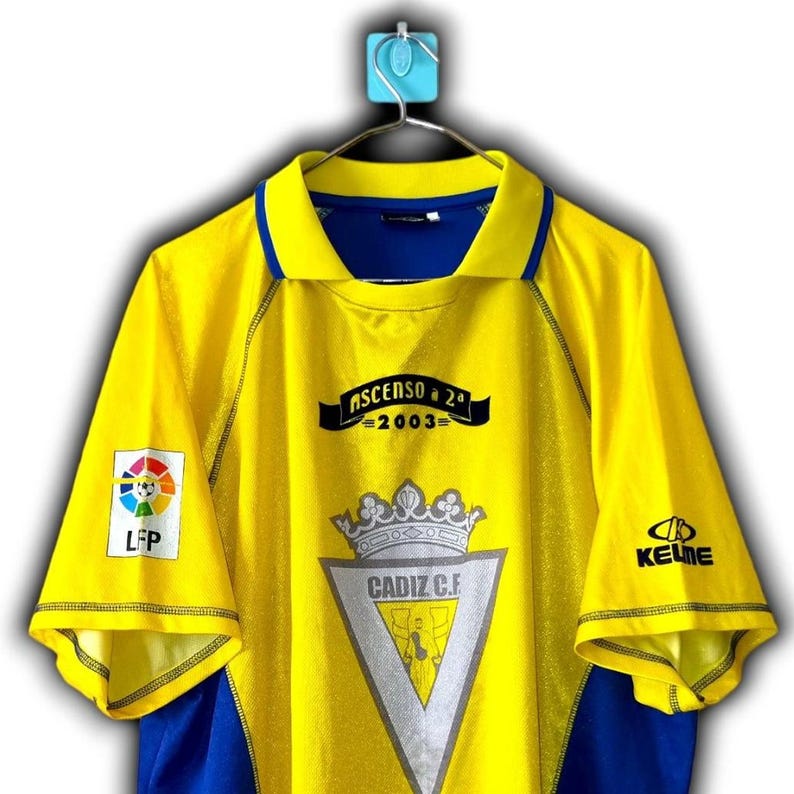 K&ouml;nnte beinhalten: Gelb-blaues Fu&szlig;balltrikot von C&aacute;diz C.F. Das Trikot tr&auml;gt den Schriftzug "ASCENSO a 2&ordf; 2003" und das Vereinswappen. Es hat auch die Logos von LFP und Kelme. Das Trikot h&auml;ngt an einem blauen Haken.
