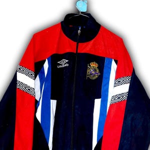 Puede incluir: Chaqueta deportiva vintage azul marino, roja y blanca con cremallera completa. La chaqueta presenta el logotipo de Umbro, un escudo con una corona y patrones geom&eacute;tricos en las mangas. La chaqueta est&aacute; colgada de una percha azul.