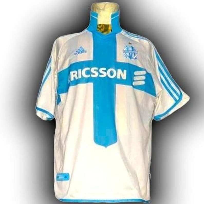 Puede incluir: Camiseta de f&uacute;tbol blanca y azul claro con el texto "RICSSON" en el pecho. La camiseta tiene un dise&ntilde;o de cruz azul en la parte delantera y rayas azules en las mangas. El logotipo de Adidas es visible.