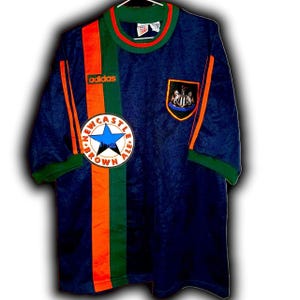 May include: A navy blue Adidas soccer jersey with orange and green accents. The jersey features the Newcastle Brown Ale logo and the Adidas logo. The jersey has short sleeves and a green collar.