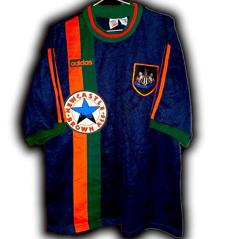 May include: A navy blue Adidas soccer jersey with orange and green accents. The jersey features the Newcastle Brown Ale logo and the Adidas logo. The jersey has short sleeves and a green collar.