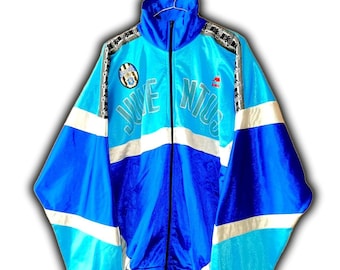 May include: A blue, white, and black Juventus track jacket. The jacket features the word "JUVENTUS" in large, white letters across the chest. The jacket has a high collar and a full-length zipper. The sleeves have white stripes and black and white patterned accents.