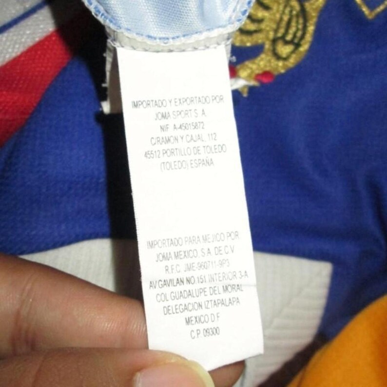 May include: A white tag with black text, featuring the words "IMPORTADO Y EXPORTADO POR JOMA SPORTS A" and other details. The tag is against a blue and yellow background.