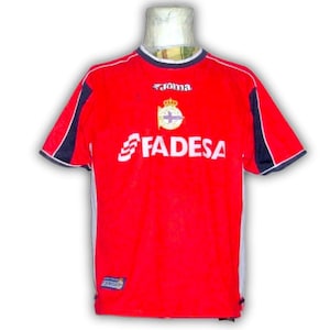 K&ouml;nnte beinhalten: Rotes Fu&szlig;balltrikot mit wei&szlig;en und marineblauen Akzenten. Das Trikot zeigt das Wort "FADESA" in Wei&szlig;, ein Teamemblem und die Marke "Joma". Es hat kurze &Auml;rmel und einen Rundhalsausschnitt.