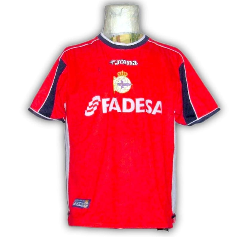 K&ouml;nnte beinhalten: Rotes Fu&szlig;balltrikot mit wei&szlig;en und marineblauen Akzenten. Das Trikot zeigt das Wort "FADESA" in Wei&szlig;, ein Teamemblem und die Marke "Joma". Es hat kurze &Auml;rmel und einen Rundhalsausschnitt.