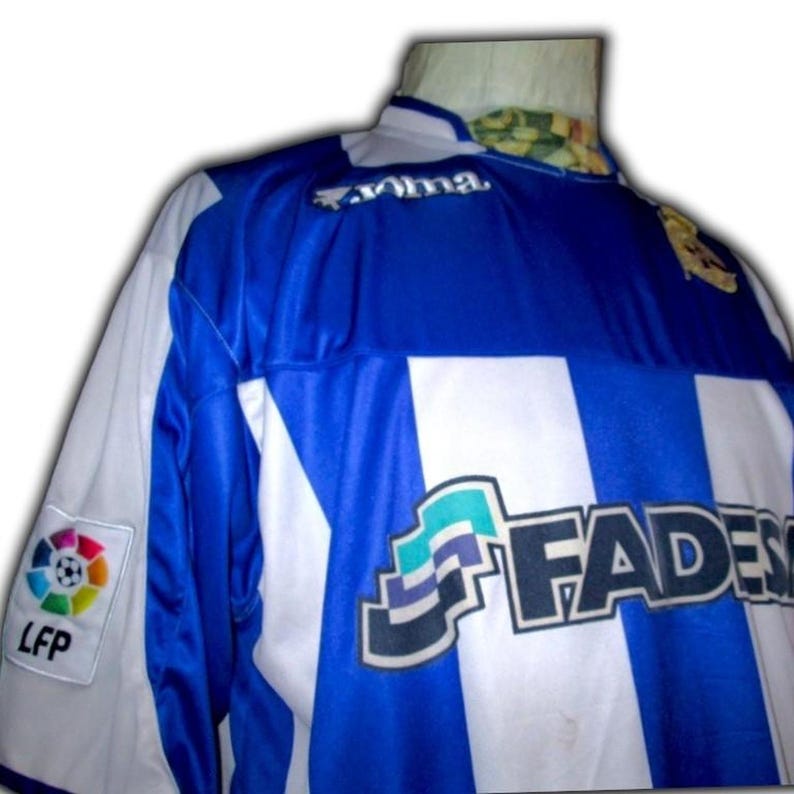 May include: A blue and white soccer jersey with the word "FADES" in large black letters. The jersey has a logo on the left sleeve and the LFP logo. The jersey has a blue collar and blue stripes.
