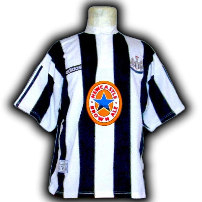 Op de afbeelding: Een zwart-wit gestreept voetbalshirt met het Adidas-logo op de schouder. Het shirt heeft een rond embleem met de tekst "Newcastle Brown Ale" en een blauwe ster. Het shirt wordt op een mannequin getoond.