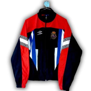 Puede incluir: Chaqueta deportiva Umbro vintage negra, roja y azul. La chaqueta tiene cremallera completa, cuello alzado y un logotipo en el pecho. Las mangas tienen patrones geom&eacute;tricos blancos y negros. Un escudo est&aacute; en el pecho.