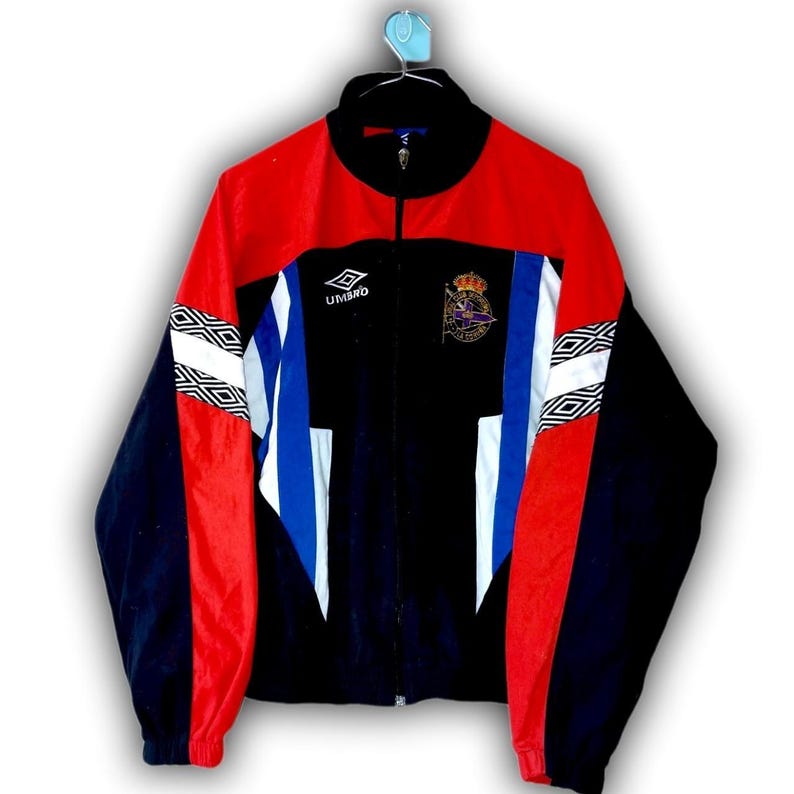 Puede incluir: Chaqueta deportiva Umbro vintage negra, roja y azul. La chaqueta tiene cremallera completa, cuello alzado y un logotipo en el pecho. Las mangas tienen patrones geom&eacute;tricos blancos y negros. Un escudo est&aacute; en el pecho.
