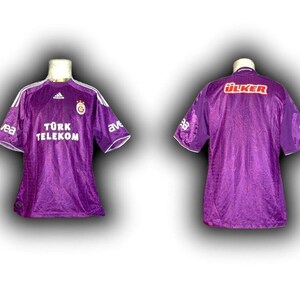 May include: A purple soccer jersey with white accents on the sleeves and collar. The front of the jersey features the text "T&Uuml;RK TELEKOM" and a team logo. The back of the jersey displays the word "&Uuml;LKER".