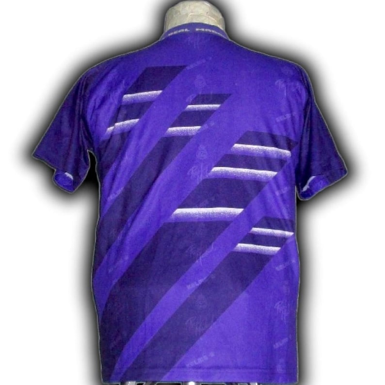 K&ouml;nnte beinhalten: Ein lila kurz&auml;rmeliges T-Shirt mit diagonalen Streifen in verschiedenen Lila- und Wei&szlig;t&ouml;nen. Das T-Shirt hat einen Rundhalsausschnitt und ein dezentes Muster. Die R&uuml;ckseite des T-Shirts ist sichtbar und zeigt das Design.