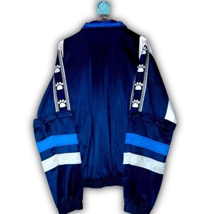 K&ouml;nnte beinhalten: Marineblaue Jacke mit wei&szlig;en und hellblauen Akzenten. Die Jacke hat wei&szlig;e Streifen mit Pfotenabdr&uuml;cken auf den &Auml;rmeln und blaue und graue Streifen an den unteren &Auml;rmeln. Sie hat einen Rei&szlig;verschluss und einen gerippten Saum.