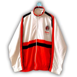 Puede incluir: Chaqueta con cremallera blanca, roja y negra con el logo del AC Milan y el texto "Motta". La chaqueta tiene cuello alto, detalles negros en los hombros y rayas rojas en los laterales. Ropa deportiva vintage.