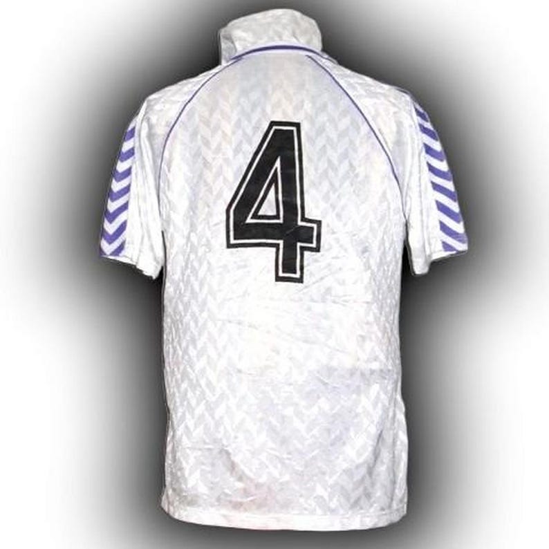 Puede incluir: Camiseta blanca de manga corta con un gran n&uacute;mero 4 negro en la espalda. La camiseta tiene un patr&oacute;n texturizado y detalles morados en el cuello y las mangas. Las mangas tienen un dise&ntilde;o a rayas en morado y blanco.