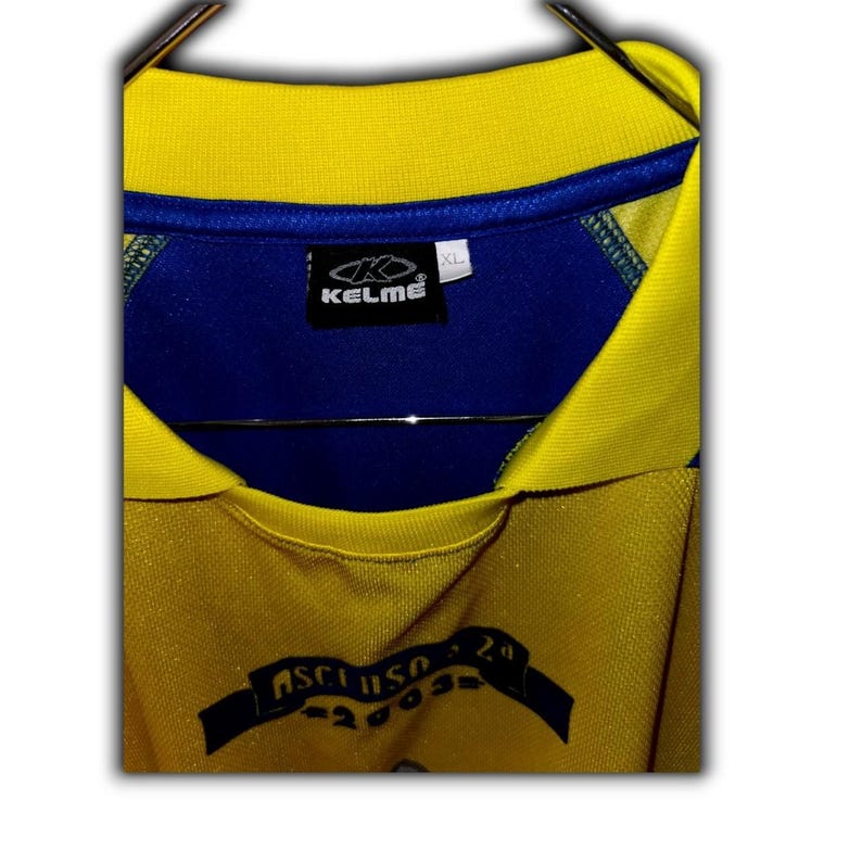 K&ouml;nnte beinhalten: Gelb-blaues XL-Fu&szlig;balltrikot mit blauem Kragen und Kelme-Logo. Das Trikot hat einen blauen Einsatz auf der oberen Brust und ein Banner mit Text am unteren Rand. Das Trikot h&auml;ngt an einem Metallb&uuml;gel.
