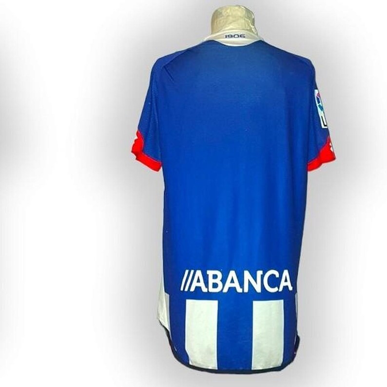 Puede incluir: Camiseta de f&uacute;tbol azul y blanca con ribetes rojos en las mangas. La camiseta muestra la palabra "ABANCA" en blanco y el n&uacute;mero "1906" en el cuello. Un logotipo es visible en la manga derecha.