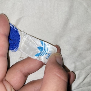 K&ouml;nnte beinhalten: Nahaufnahme eines blau-wei&szlig;en Adidas-Etiketts mit dem Adidas-Trefoil-Logo in Blau. Das Etikett ist an einem blauen Kleidungsst&uuml;ck mit wei&szlig;er Bord&uuml;re befestigt. Der Hintergrund ist ein wei&szlig;er Stoff.