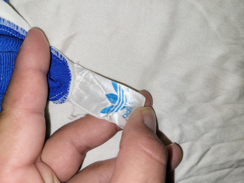 K&ouml;nnte beinhalten: Nahaufnahme eines blau-wei&szlig;en Adidas-Etiketts mit dem Adidas-Trefoil-Logo in Blau. Das Etikett ist an einem blauen Kleidungsst&uuml;ck mit wei&szlig;er Bord&uuml;re befestigt. Der Hintergrund ist ein wei&szlig;er Stoff.