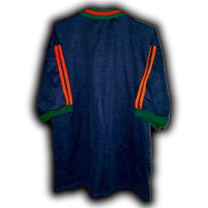 May include: Navy blue jersey with orange stripes on the sleeves and a green collar and sleeve cuffs. The jersey has a textured pattern and a crew neck with orange and green trim.