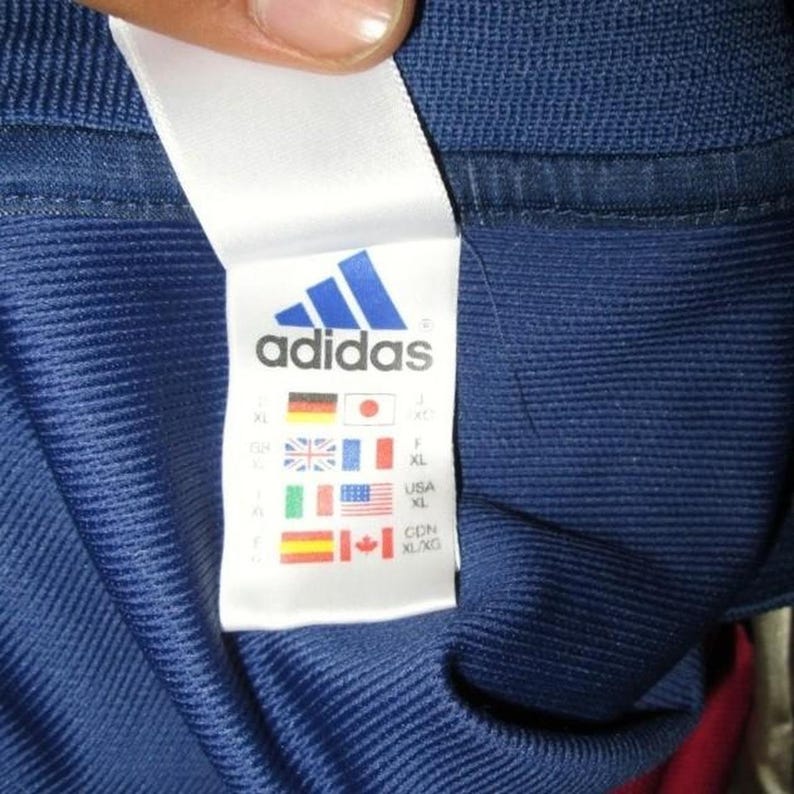 May include: Close-up of a blue Adidas tag. The tag features the Adidas logo and flags representing various countries, including Germany, Japan, the UK, France, the USA, and Canada. The tag also indicates the size as XL.