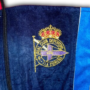 K&ouml;nnte beinhalten: Nahaufnahme einer marineblauen Jacke mit einem goldgestickten Emblem. Das Emblem zeigt eine Krone, ein Kreuz und die Worte "REAL CLUB DEPORTIVO LA CORUNA" in goldener Schrift. Die Jacke hat einen blauen und roten Streifen.