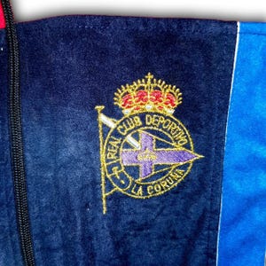 Puede incluir: Primer plano de una chaqueta azul marino con un emblema bordado en oro. El emblema presenta una corona, una cruz y las palabras "REAL CLUB DEPORTIVO LA CORUNA" en letras doradas. La chaqueta tiene una raya azul y roja.