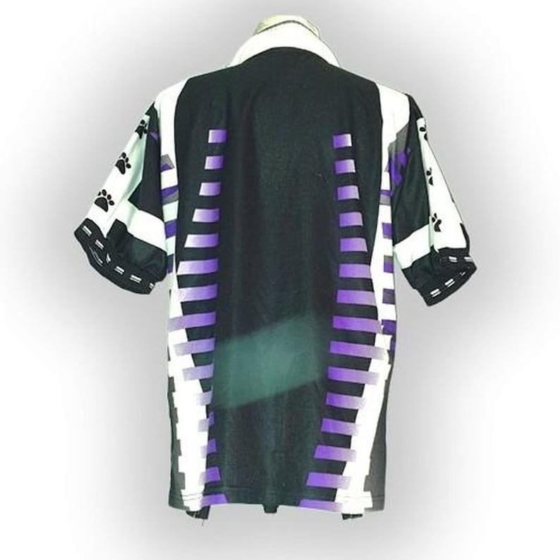May include: Black and white short-sleeved shirt with purple accents. The shirt features a collar and a pattern of paw prints on the sleeves. Vertical purple stripes run down the center of the shirt, with white and black geometric designs.