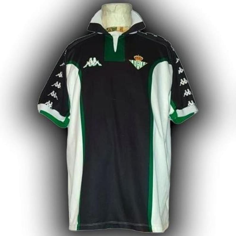 Op de afbeelding: Een zwart, wit en groen voetbalshirt met een kraag. Het shirt heeft het Kappa-logo op de mouwen en de borst, samen met een teamembleem. Het ontwerp omvat witte zijpanelen en groene afwerking.