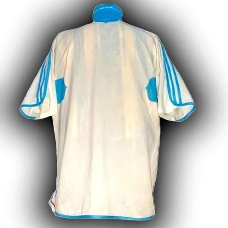 Puede incluir: Camiseta deportiva color crema con detalles en azul claro. La camiseta presenta rayas azules claras en las mangas y un cuello azul claro. La parte trasera de la camiseta es visible.