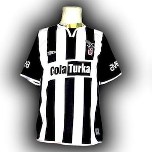 May include: A black and white striped soccer jersey with short sleeves. The jersey has a V-neck collar and the text "cola Turka" across the chest. The sleeves are black with white trim, and there is a team logo on the upper chest.