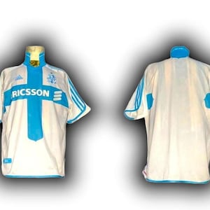 Puede incluir: Una camiseta de f&uacute;tbol blanca con detalles en azul claro. La camiseta presenta un dise&ntilde;o de cruz azul claro en la parte delantera, con la palabra "RICSSON" impresa en el centro. El logo de Adidas es visible en la parte superior izquierda.