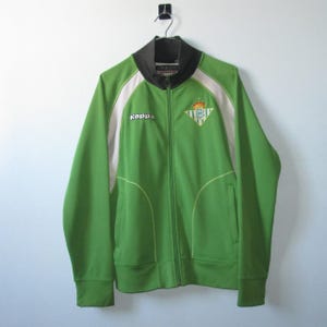 K&ouml;nnte beinhalten: Gr&uuml;ne Trainingsjacke mit Rei&szlig;verschluss und wei&szlig;en und schwarzen Akzenten. Die Jacke hat das Kappa-Logo und ein Team-Emblem auf der Brust. Sie h&auml;ngt an einem schwarzen Haken vor einer wei&szlig;en Wand.