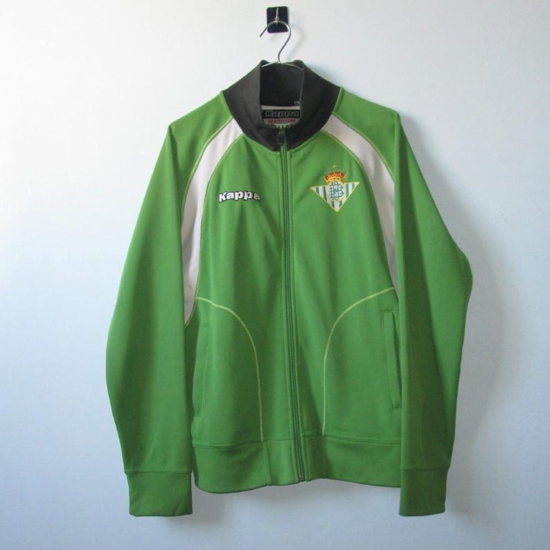 K&ouml;nnte beinhalten: Gr&uuml;ne Trainingsjacke mit Rei&szlig;verschluss und wei&szlig;en und schwarzen Akzenten. Die Jacke hat das Kappa-Logo und ein Team-Emblem auf der Brust. Sie h&auml;ngt an einem schwarzen Haken vor einer wei&szlig;en Wand.