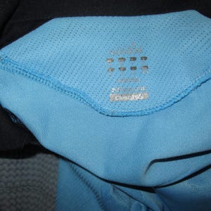 Puede incluir: Primer plano del cuello de una camiseta deportiva azul claro. El cuello presenta una tela de malla y una etiqueta con el texto "Clima365". La camiseta est&aacute; hecha de un material transpirable, adecuado para deportes y ejercicio.