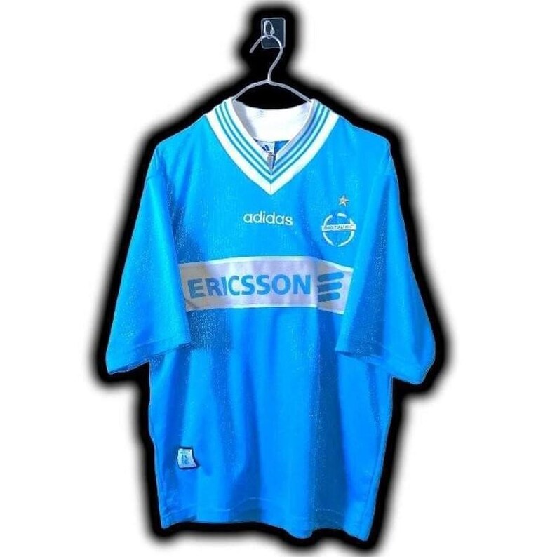 Puede incluir: Camiseta de f&uacute;tbol vintage azul claro con detalles blancos y azul marino. Presenta el logo de Adidas, el patrocinador Ericsson y un emblema del equipo. La camiseta tiene cuello en V y mangas cortas.