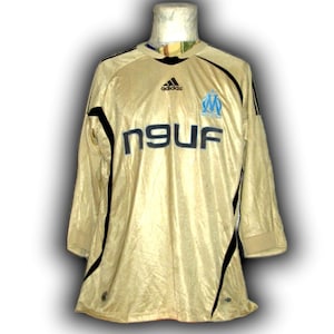 K&ouml;nnte beinhalten: Ein goldfarbenes Fu&szlig;balltrikot mit langen &Auml;rmeln, schwarzen Akzenten und dem Adidas-Logo. Das Trikot zeigt den Text "n9uf" in Schwarz und ein hellblaues Emblem. Das Trikot befindet sich auf einer Schaufensterpuppe.