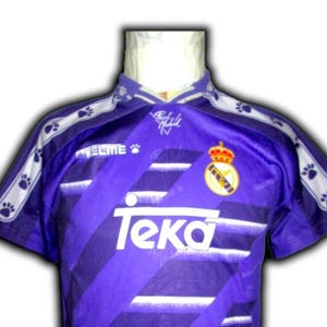 K&ouml;nnte beinhalten: Violettes Fu&szlig;balltrikot mit wei&szlig;en diagonalen Streifen und dem Teka-Logo. Das Trikot hat einen Kragen und wei&szlig;e Pfotenabdruck-Akzente auf den Schultern. Das Real Madrid-Wappen befindet sich auf der Brust.