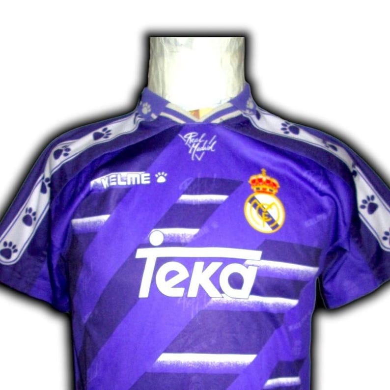 K&ouml;nnte beinhalten: Violettes Fu&szlig;balltrikot mit wei&szlig;en diagonalen Streifen und dem Teka-Logo. Das Trikot hat einen Kragen und wei&szlig;e Pfotenabdruck-Akzente auf den Schultern. Das Real Madrid-Wappen befindet sich auf der Brust.