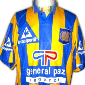 May include: A vintage soccer jersey in blue and yellow, featuring a collared neckline and short sleeves. The jersey has a vertical blue stripe down the center and the text "general paz seguros". The Le Coq Sportif logo is visible.